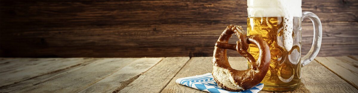 A frothy glass of beer sits on a wooden table next to a pretzel on a blue and white napkin, capturing the spirit of Simi Valley Oktoberfest.