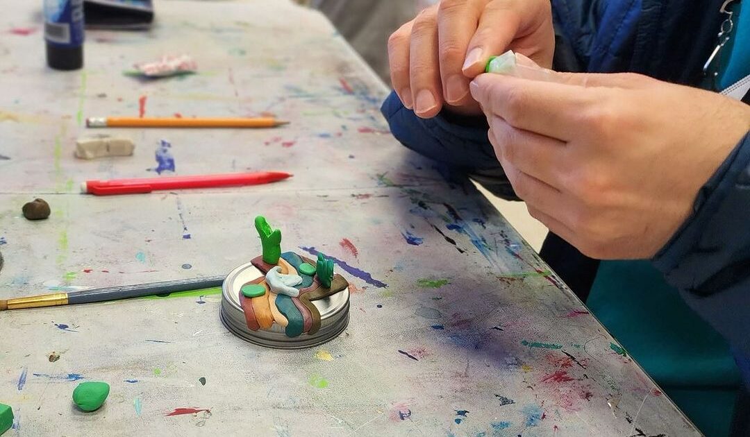 Person shaping green clay with hands at a messy table, surrounded by art supplies like paintbrushes and markers.