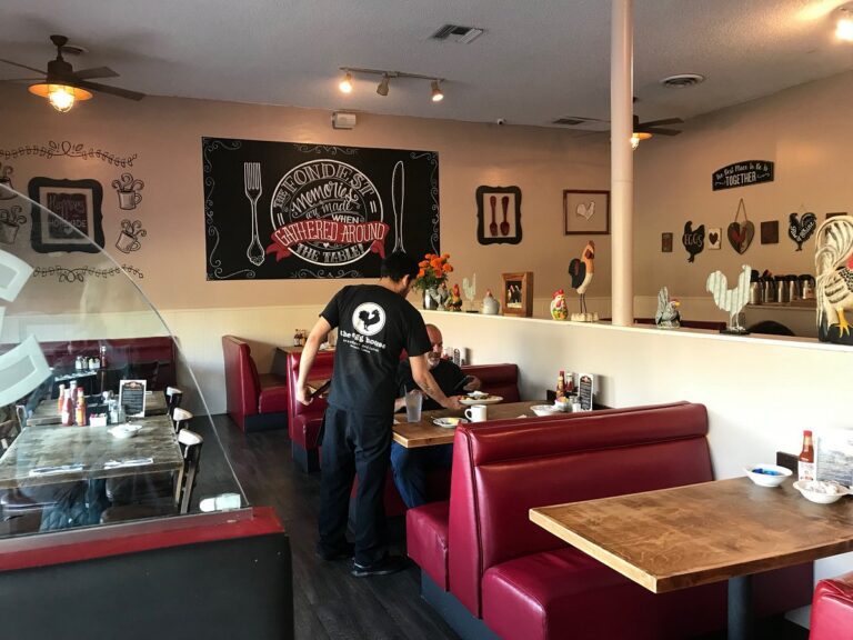 A waiter serves a customer at a diner with red booth seating and wall decorations, including text and kitchen-themed art.