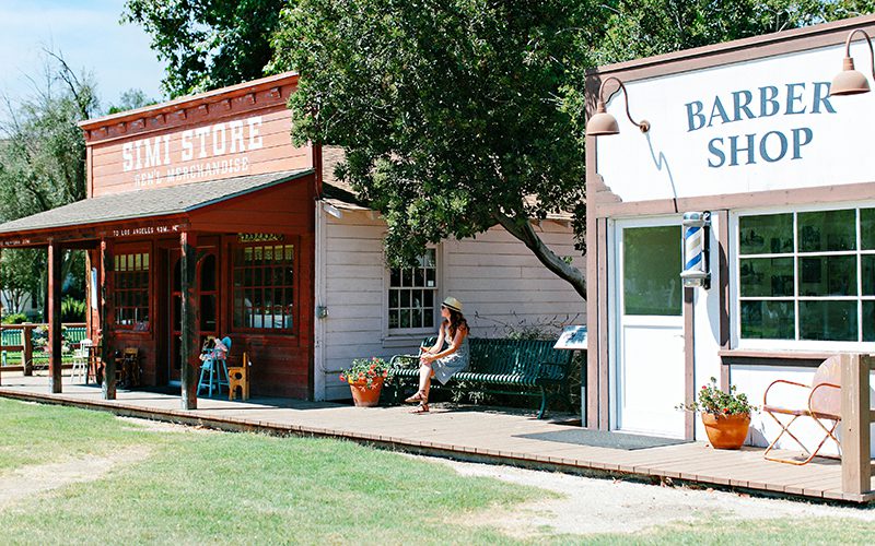 A woman sits on a bench near a vintage-style general store and barber shop on a wooden sidewalk, soaking in the charm of one of the many things to do in Simi Valley.