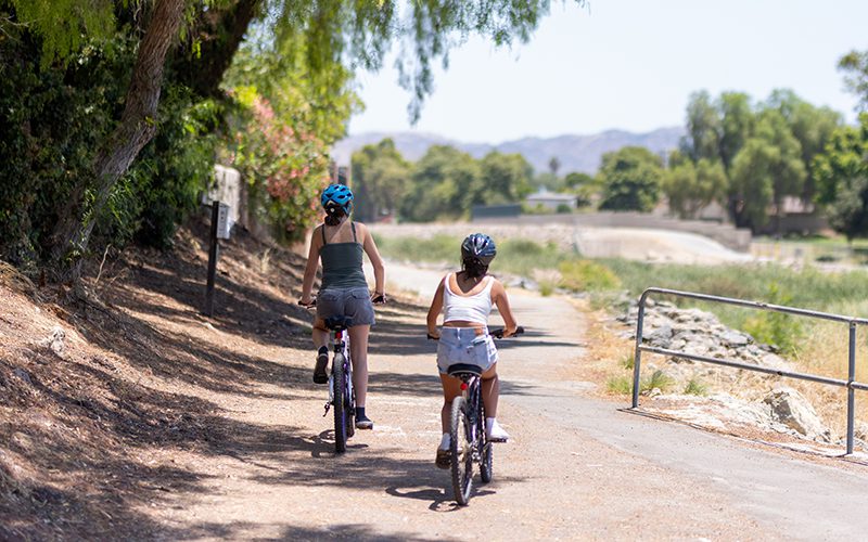 Two people wearing helmets ride bicycles on a sunny path with trees and a fence nearby, enjoying one of the many relaxing things to do in Simi Valley.