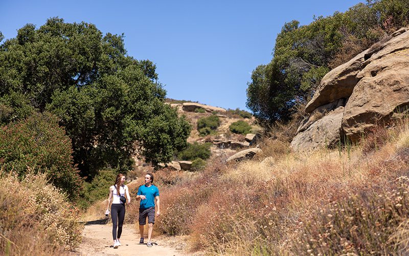 Taking in one of the many things to do in Simi Valley, two people walk on a dirt path surrounded by trees and rocks under a clear blue sky.