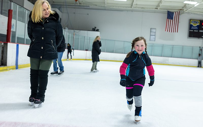 A woman and a smiling child find joy ice skating indoors, one of the delightful things to do in Simi Valley. The U.S. flag adds a touch of pride to the scene as they glide gracefully across the rink.