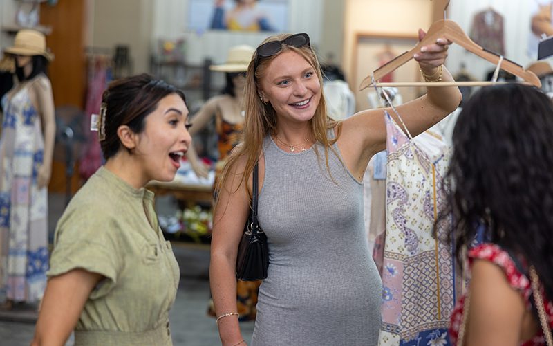 Three women shopping in Simi Valley, one holding up a dress on a hanger, all smiling and admiring it. Clothing racks and colorful dresses fill the background, making it a delightful addition to your list of things to do in Simi Valley.