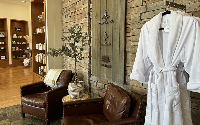 The spa waiting area in Simi Valley offers two inviting brown leather chairs, a plush white robe hanging nearby, and a small side table adorned with a vibrant plant. Shelves stocked with premium products complete this tranquil spot, creating one of the go-to things to do in Simi Valley for relaxation enthusiasts.