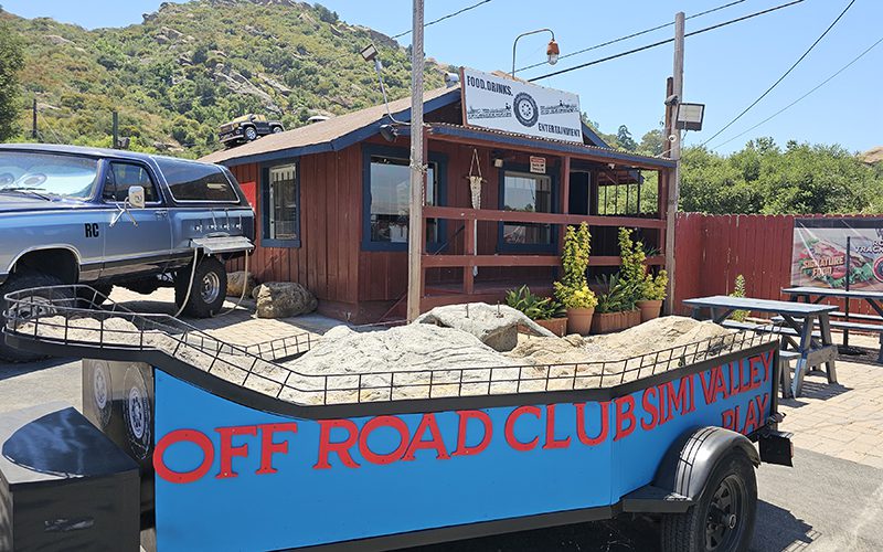 A blue trailer with "Off Road Club Simi Valley" is parked in front of a rustic building labeled "Food Service." Nearby, a classic car adds charm, with mountains gracing the backdrop—one of the many things to do in Simi Valley for adventure and nostalgia lovers alike.