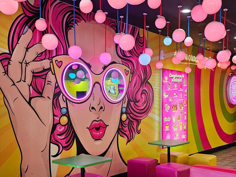 Colorful interior with pop art wall mural of a woman in cat-eye sunglasses. Pink and white ball lights hang from the ceiling. Bright furniture and a vibrant menu add to the lively atmosphere.