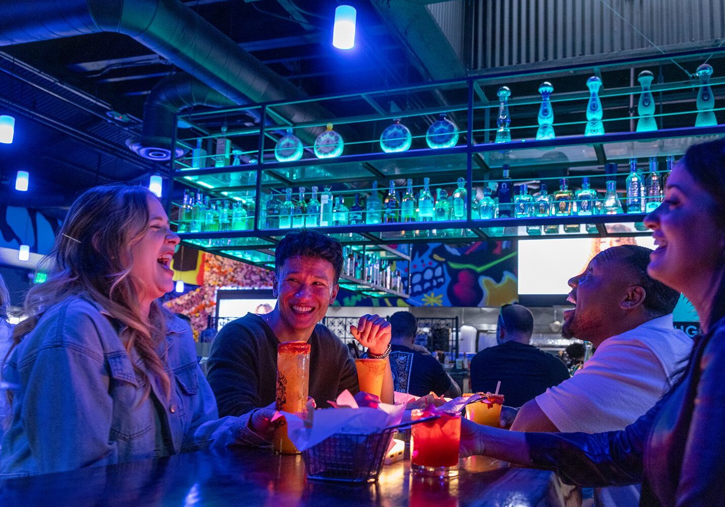 People laughing and enjoying drinks at a vibrant bar under blue and purple lighting in Simi Valley, with colorful decor and shelves of bottles creating a lively backdrop—one of the many fun things to do in Simi Valley.